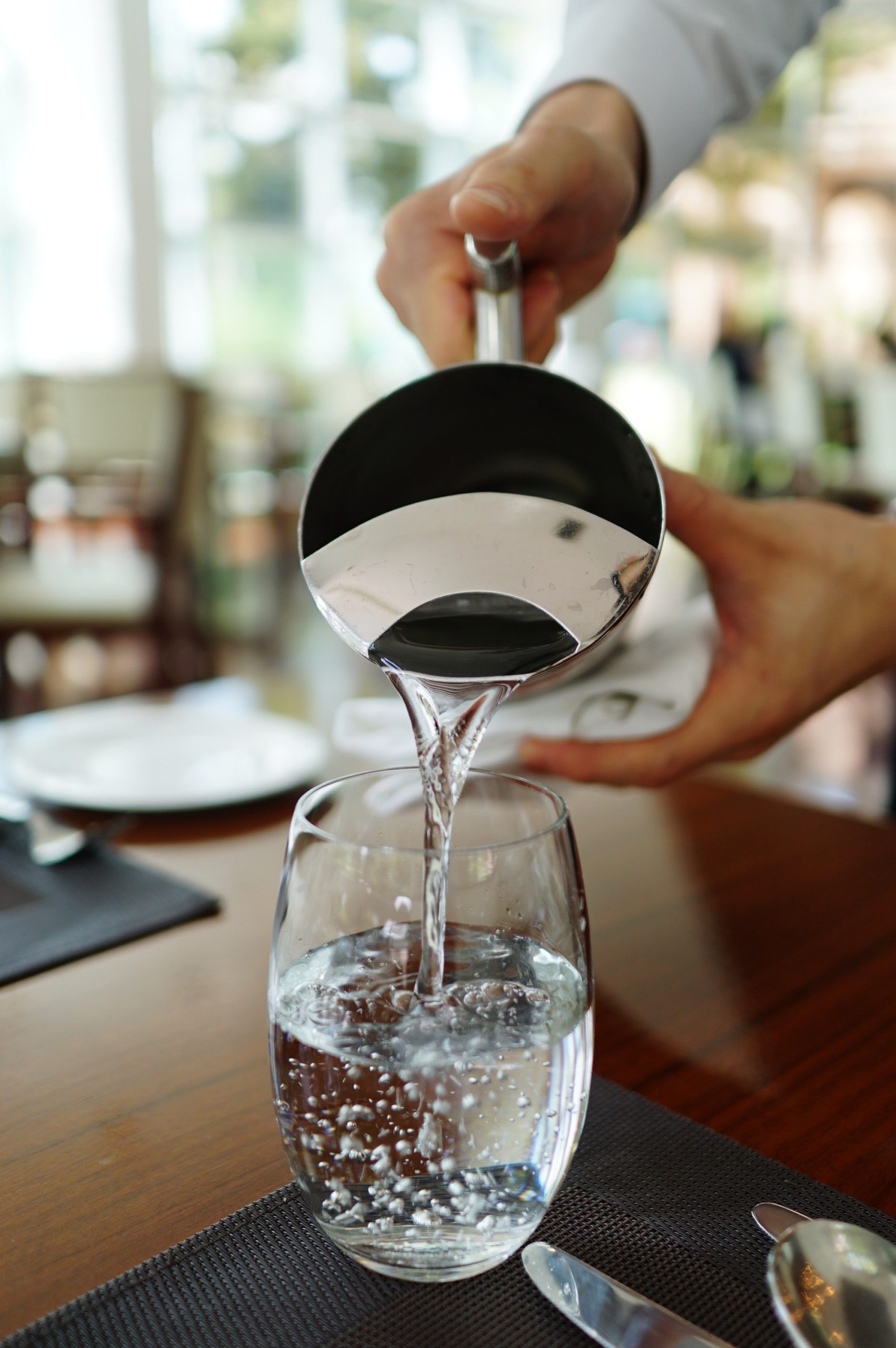 Hands pouring water from a steel pitcher into a glass on a table set for a meal. Blurred background