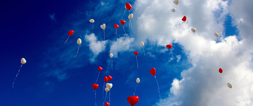 Many red and white heart-shaped balloons released and flying into a blue sky with white clouds