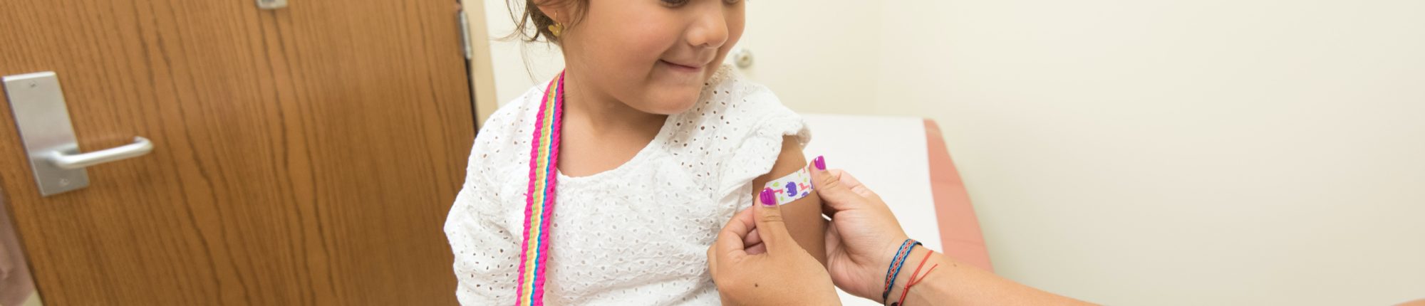 a young girl sits on a doctor's office bed. A person puts a bandage on her arm