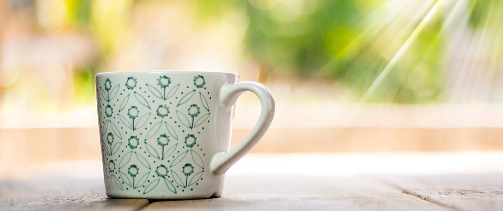Close-up of a white mug with a green floral pattern sitting on a wooden table in front of a window