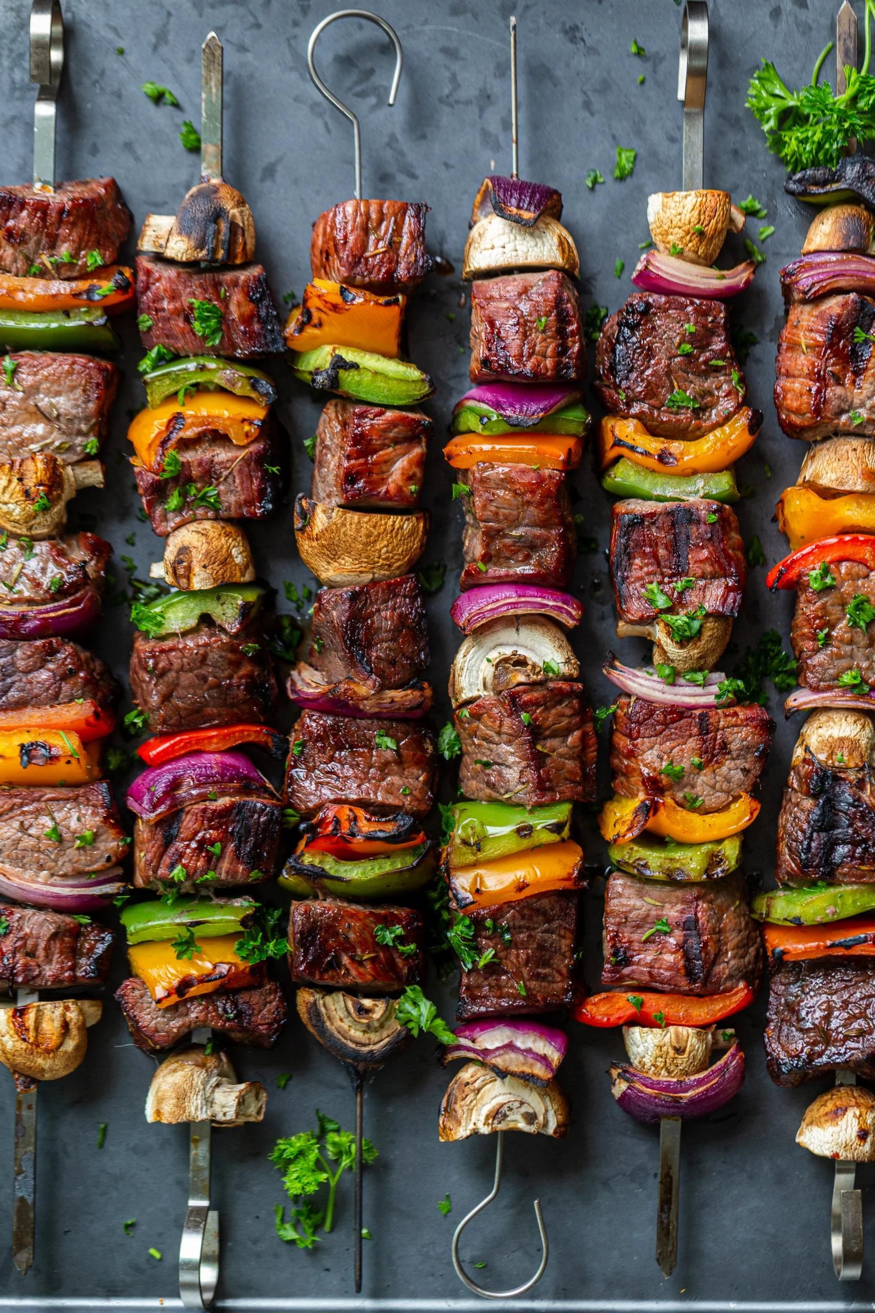 Six metal skewers side-by-side filled with chunks of cooked meat, mushrooms, peppers, and onion