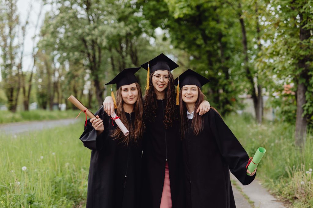 Three light-skinned women in black graduation caps and gowns pose together with their diplomas