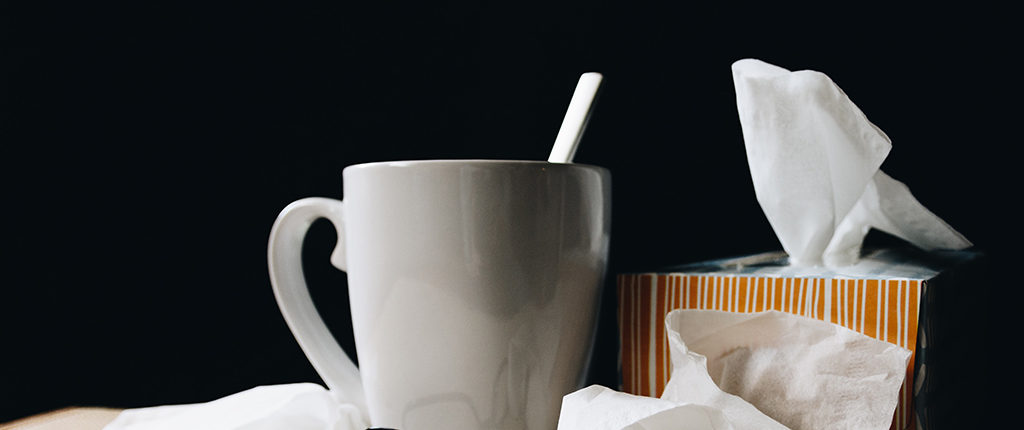A gray mug with a spoon, used tissues, and a tissue box against a black background