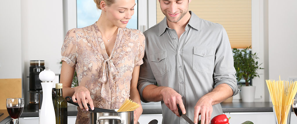 Pale-skinned man and woman stand at a kitchen counter cooking pasta and chopping vegetables