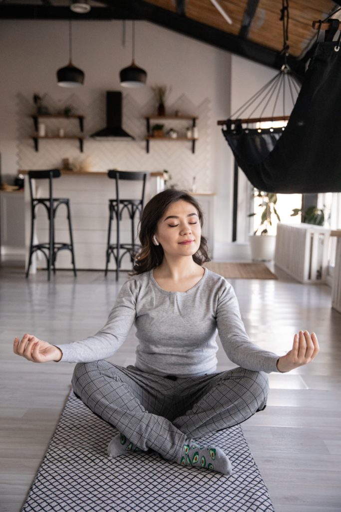 Pale-skinned woman sits crosslegged with arms on knees and eyes closed, wearing comfy gray clothes