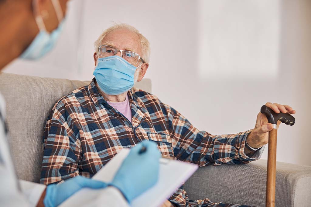 Masked older male patient with cane sits on a couch inside home beside masked doctor with clipboard