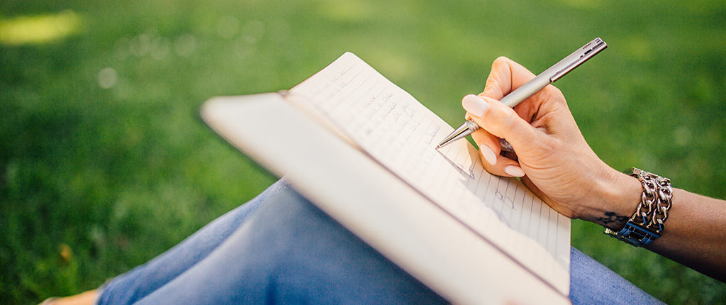 Close-up of hands writing with a pen in a journal propped on bent knees. The writer sits on grass