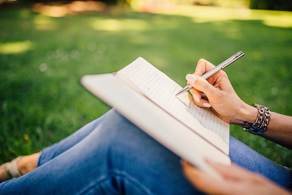 Close-up of hands writing with a pen in a journal propped on bent knees. The writer sits on grass