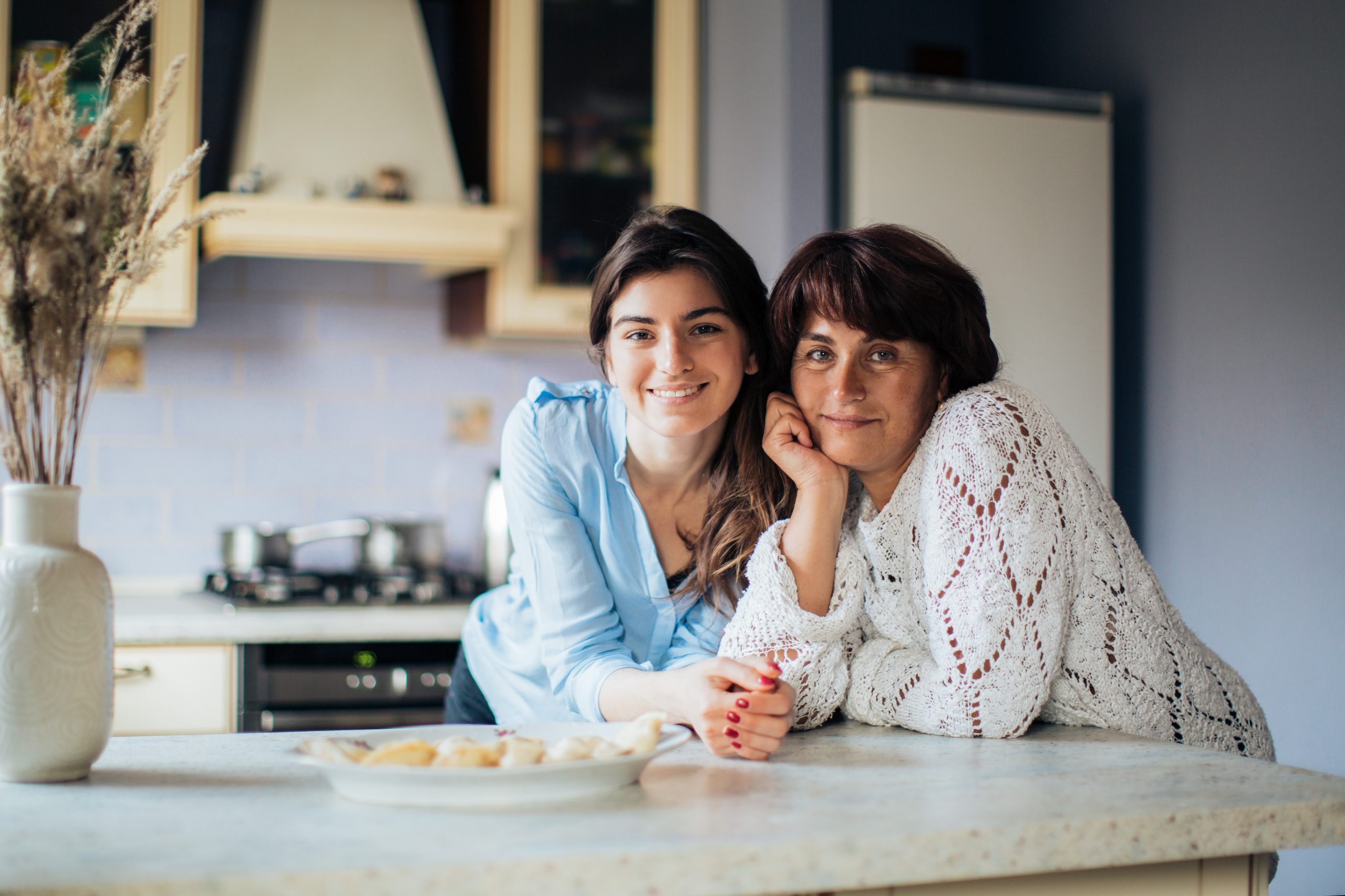 A middle-aged woman and a young woman both lean on a countertop in a home kitchen