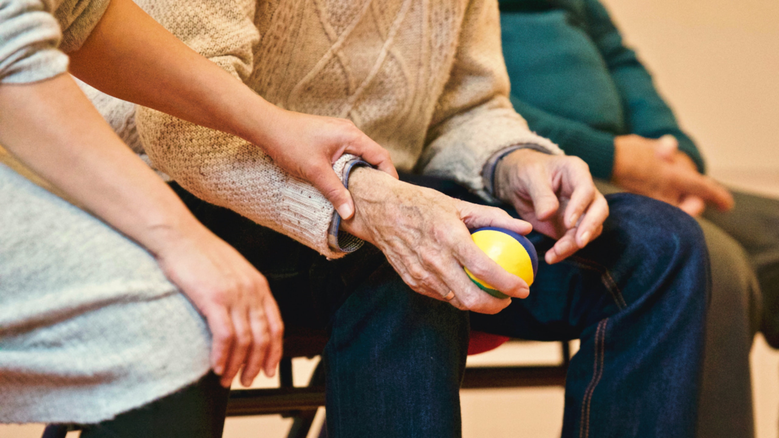 An older man in a sweater holds a ball. A woman in the foreground puts her hand on his wrist.