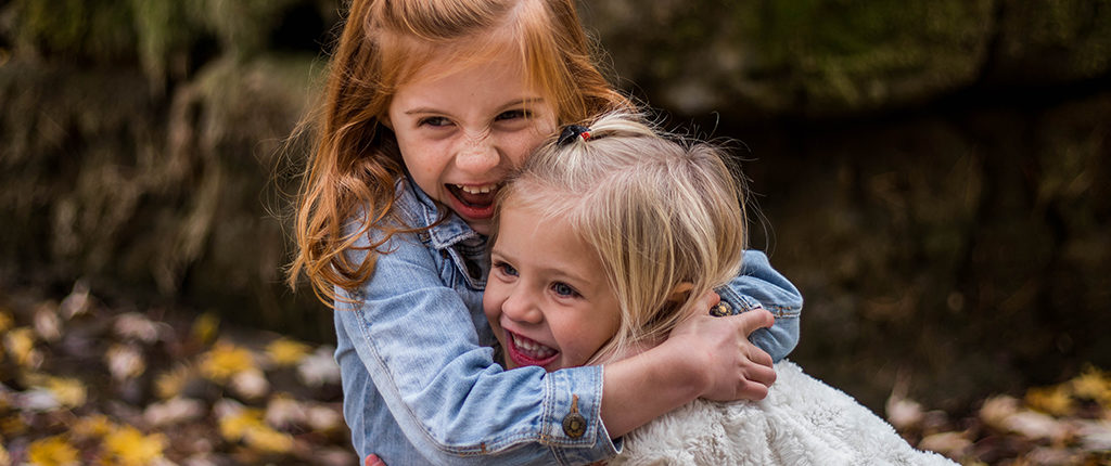 Two young girls in light jackets hug each other as they smile, blurred background of fallen leaves