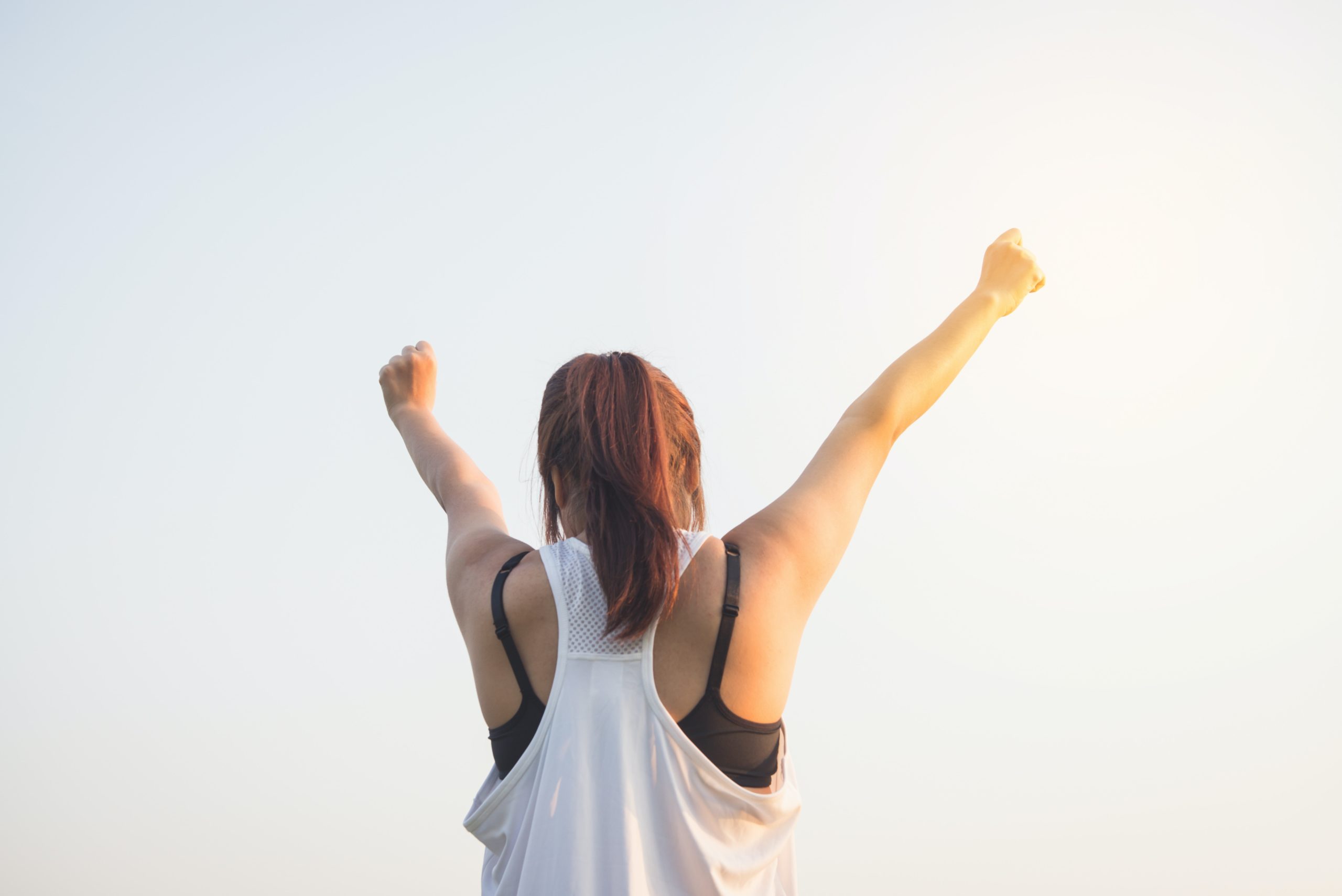 Woman with pale skin from the waist up, pictured from behind, arms raised to the sky in victory