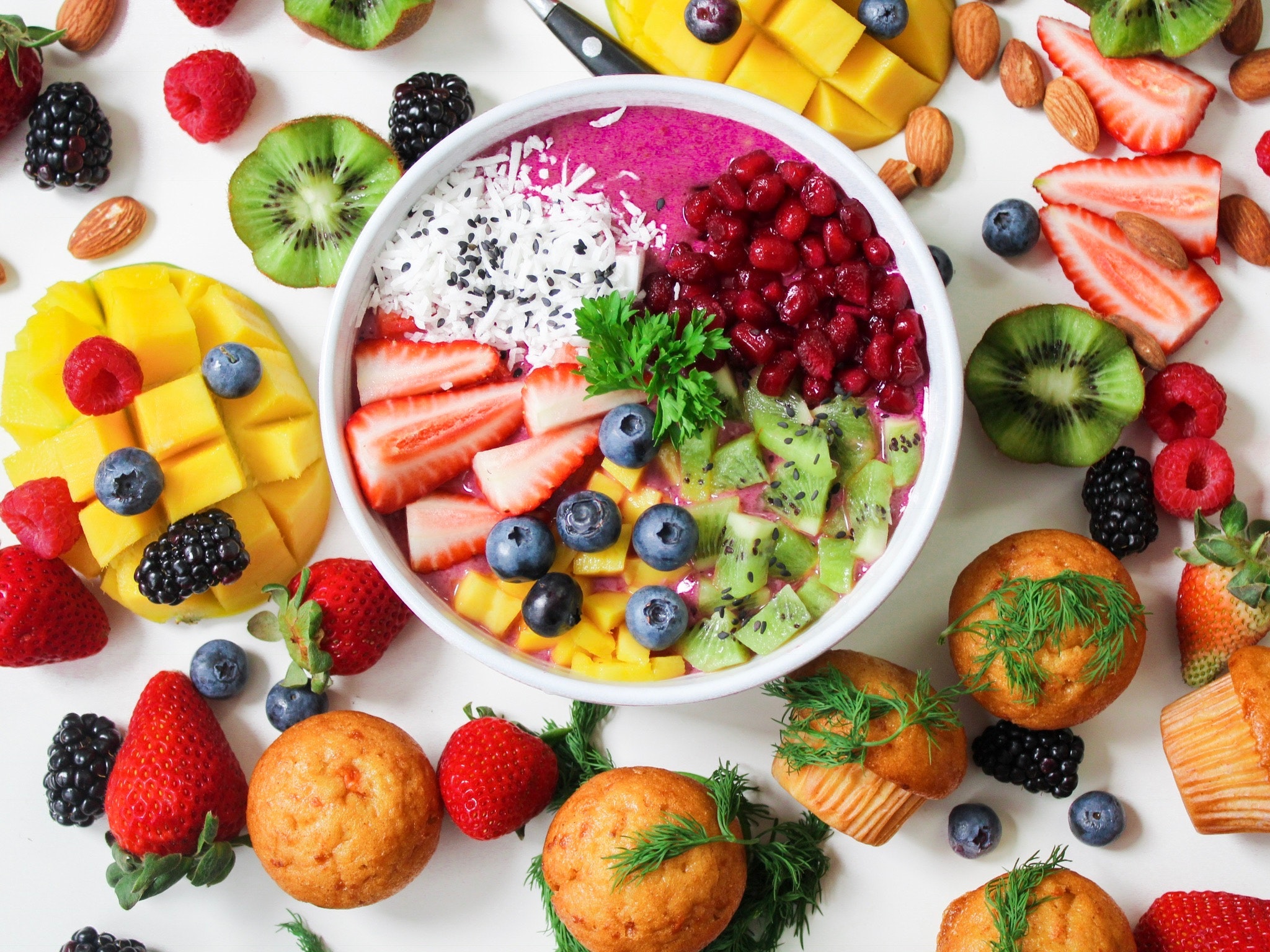 Top-down, a bowl of cut-up fruit sits on a white table, surrounded by cut fruit and several muffins