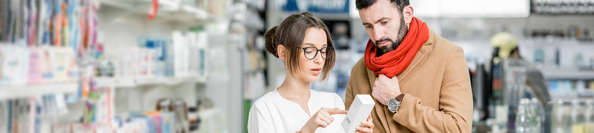 Female pharmacist is pointing at box of medicine next to male with coat and scarf on in pharmacy