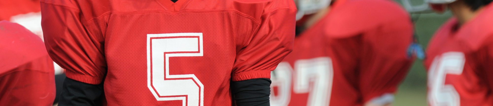 Close-up of American football player's chest in a red jersey with a white five, more players behind