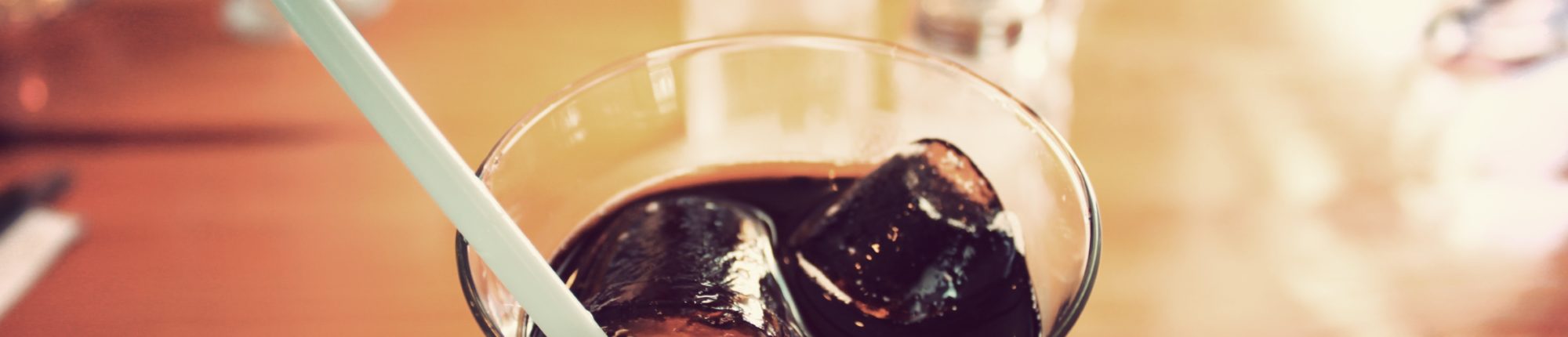 Extreme close-up of the top of a glass with brown soda, ice, and a straw in it, blurred background