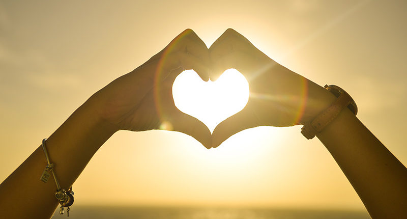 A woman makes a heart shape with her hands in front of a sunset