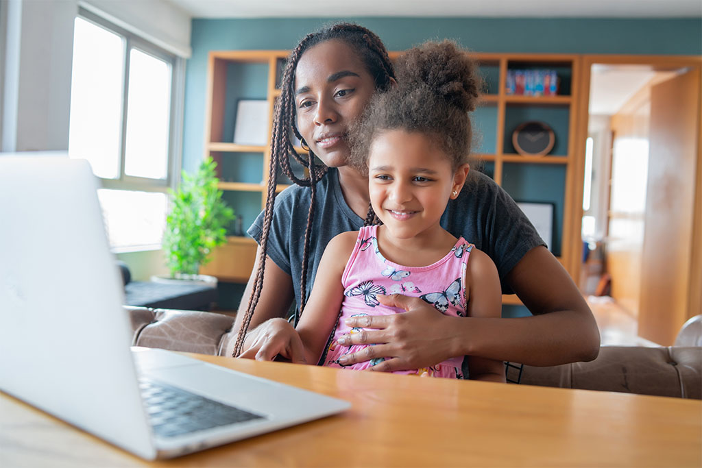 Woman holding a child in her lap in a living room while they both view a telehealth call on a laptop