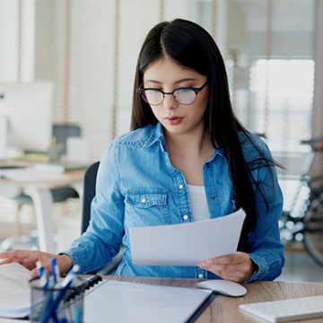 Young woman with glasses is sitting at desk in building and reviewing paperwork