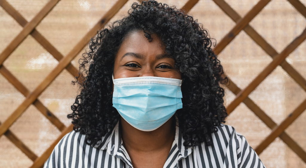 Portrait of masked woman, standing outside and smiling at the camera on a warm, bright day. 