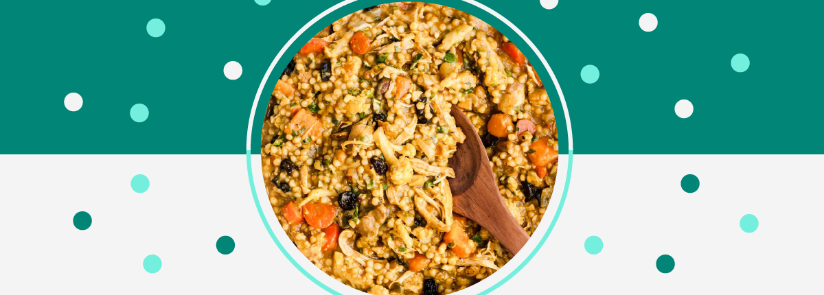 Bowl of cooked chicken, couscous, carrots and cherrries. A wooden spoon is inserted into the stew