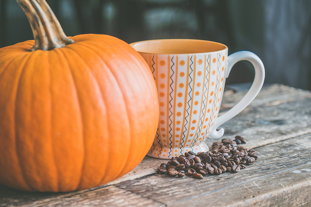 A pumpkin sits next to a coffee mug with a small pile of beans in the foreground