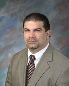 Portrait of Joe Montone wearing a light gray top, white collared shirt, and checkered tie