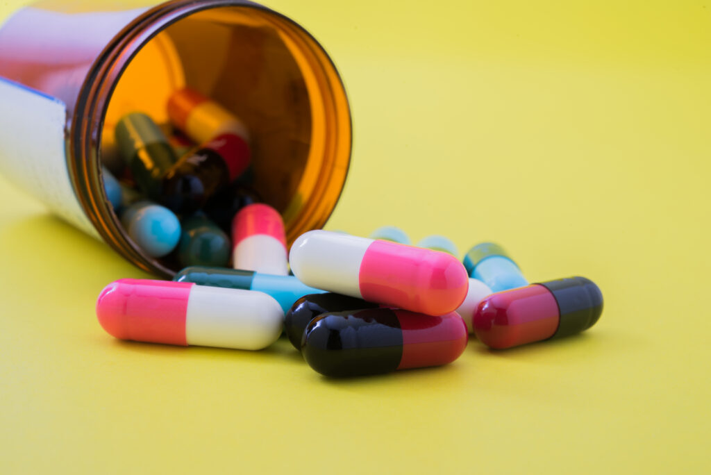 Colorful assorted medication capsules spilling from an amber prescription bottle on yellow background.