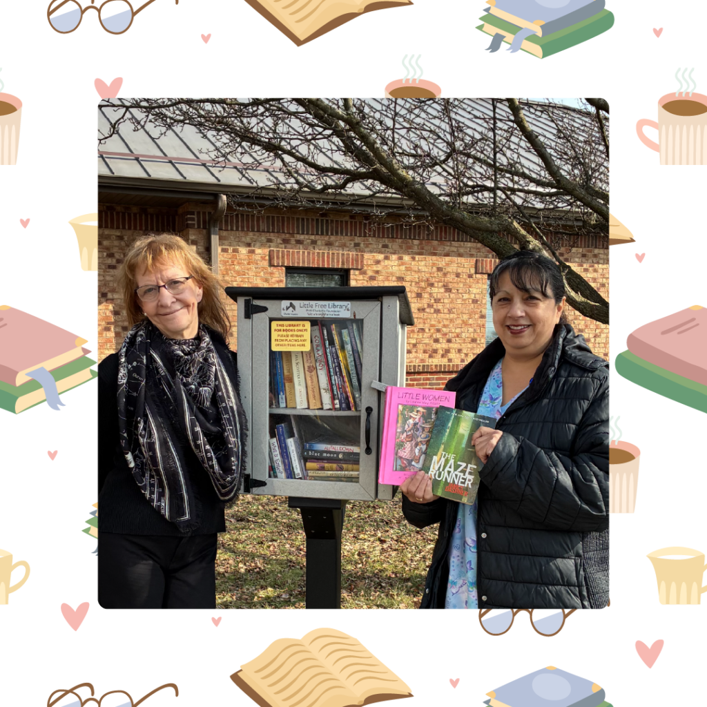 Two women stand beside a Little Free Library box filled with books outdoors on a sunny day.