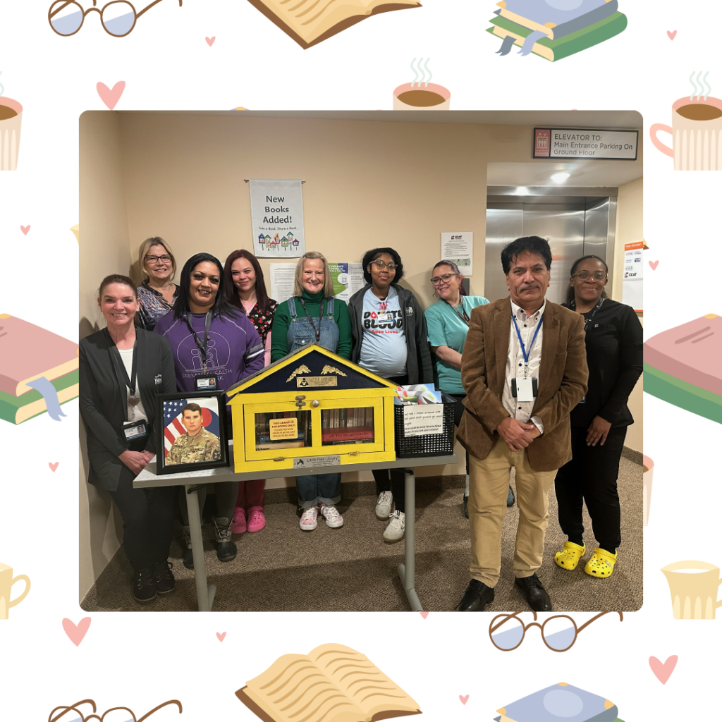 Group of people standing behind a table displaying a yellow miniature library and a framed military photo indoors.