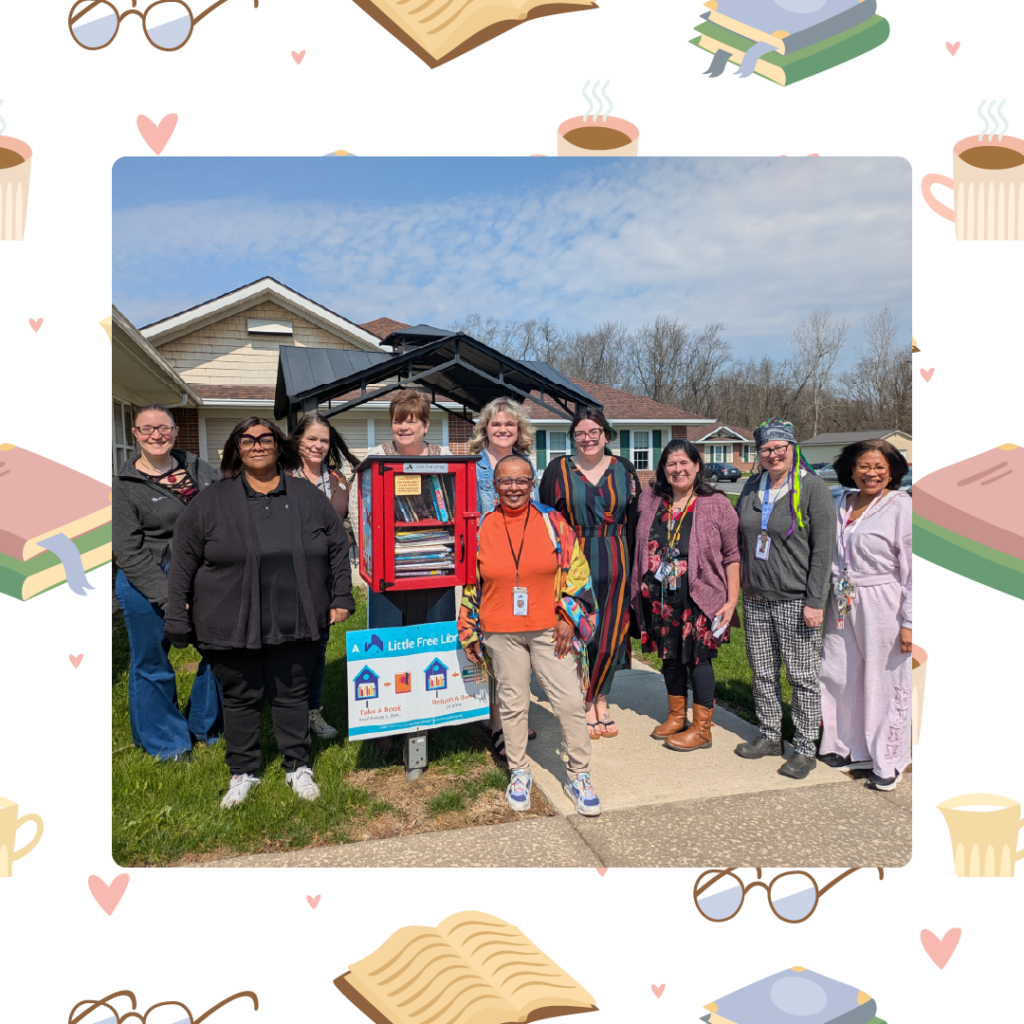 Group of adults standing outdoors around a Little Free Library box filled with books on a sunny day.