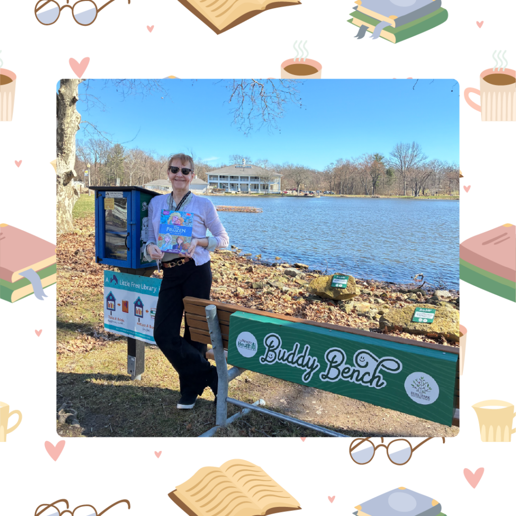 Person standing by a small outdoor library box near a lake, holding a children's book.